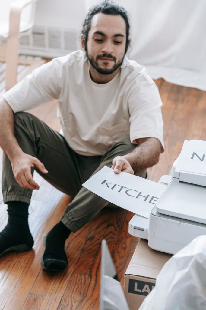 A young man sits on the floor labeling boxes for moving in a sunlit room.
